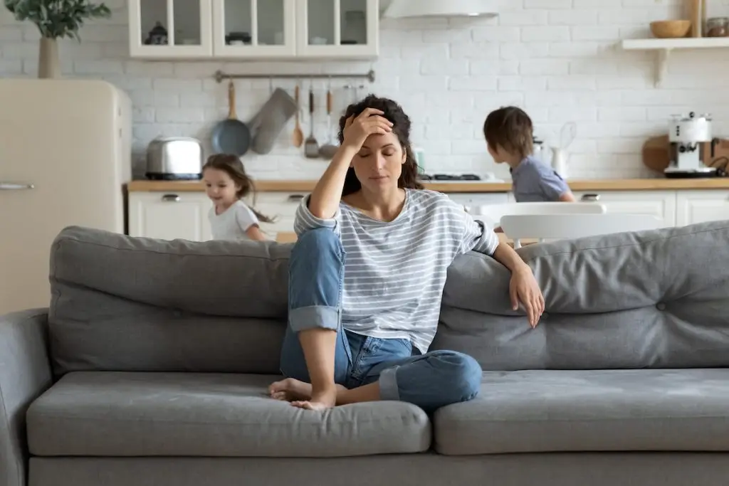 Women sitting on sofa with hand on forehead while kids run wildly behind her