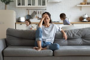 Women sitting on sofa with hand on forehead while kids run wildly behind her